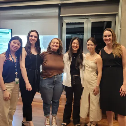 Seis mulheres sorrindo diante de um totem com pôster