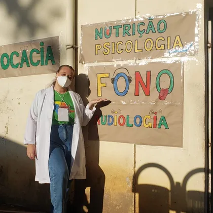 Luciene apontando uma placa pintada “Fono / Audiologia”